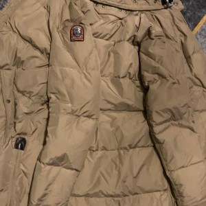 Beige dunjacka från Parajumpers M - Säljer en beige Parajumpers dunjacka i storlek medium. Jackan har klassisk puffer-design, patch på ärmen och detaljer som robust dragkedja och hög krage. Perfekt för kalla vinterdagar och riktigt snygg till streetwear-stil.