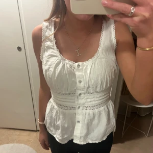 Vit ärmlös blus med spetsdetaljer - Supersöt vit blus i bomull med knappar framtill, spetsdetaljer och volang nedtill. Den är från hm i storlek xs, men lappen är bortklippt!