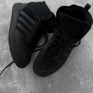 Svarta höga sneakers från Adidas - Säljer ett par svarta höga sneakers från Adidas med klassiska tre ränder på sidan. Skorna har snörning, rund tå Perfekta för dig som gillar en clean och stilren look. Använd 2 tillfällen då det var fel strl för mig  fodrade och de är i mkt bra skick se sulan 