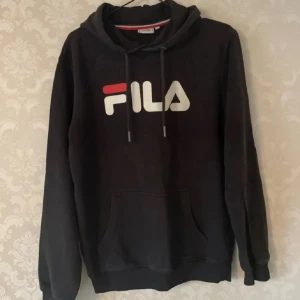 Svart hoodie från FILA med logga - Svart hoodie från FILA i storlek XS med stor vit och röd logga framtill. Tröjan har huva med snörning och en klassisk känguruficka. Perfekt för dig som gillar sportig och avslappnad stil.