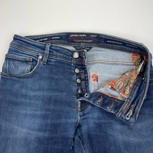Jacob Cohen jeans 622 - Snygga blå jeans från Jacob Cohen med klassisk femficksdesign och knäppning med knappar. Jeansen har en cool detalj med färgglatt mönstrat tyg på insidan och en beige patch med logga bak. Perfekt passform och stilren look.