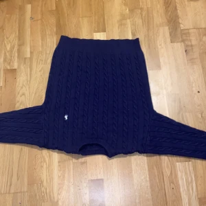 Mörkblå kabelstickad tröja Polo Ralph Lauren - Mörkblå kabelstickad tröja från Polo Ralph Lauren med klassisk rund halsringning och vit broderad logga på bröstet. Tröjan har lång ärm och ribbade muddar, perfekt för lager-på-lager under kyliga dagar.