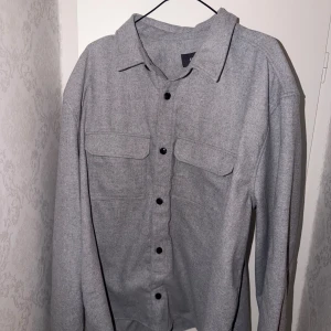 Grå overshirt - Stilren grå overshirt från H&M i storlek XXL. Skjortan har klassisk krage, svarta knappar framtill och två stora bröstfickor med lock. Perfekt lager-på-lager-plagg med lång ärm och rak passform. Helt ny och oanvänd