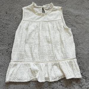 Kräm vit blus - Säljer en söt vit blus med ärmlös design och volangkant nedtill. Toppen har broderade spetsdetaljer upptill.