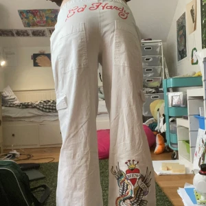 Vita cargobyxor från Ed Hardy - Säljer ett par vita cargobyxor från Ed Hardy med snörning i midjan och coolt New York-tryck på benet. Byxorna har bred passform, stora fickor på sidorna och röd Ed Hardy-logga bak och fram. Perfekta för dig som gillar streetstyle och detaljer. Liten fläck på framsidan går att tvätta bort.