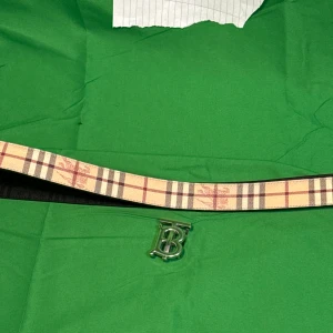 Burberry bälte med klassiskt rutmönster - Snyggt Burberry bälte med den ikoniska beigea och svarta rutan. Bältet har en silverfärgad metallspänne med ett stort B framtill. Ena sidan är svart med diskret mönster, andra sidan har klassiskt rutigt Burberry-tryck. Perfekt accessoar för att lyfta din outfit.Liknande modell som originalet!