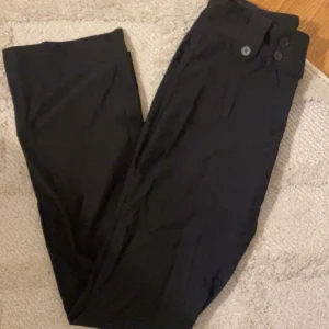 Svarta kostymbyxor med knappar - Snygga svarta kostymbyxor med raka ben och flera dekorativa knappar i midjan både fram och bak. Aldrig använda