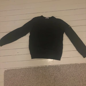 Svart sweatshirt från Stone Island - Svart sweatshirt från Stone Island med klassisk rund hals och ikonisk logotyp-patch på vänster ärm. Tröjan har ribbade muddar och är tillverkad i mjuk bomull, perfekt för en clean och stilren look.