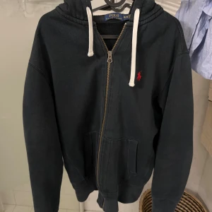 Svart hoodie från Polo Ralph Lauren - Svart hoodie från Polo Ralph Lauren med dragkedja, vit snörning i huvan och röd broderad logga på bröstet. Tröjan har känguruficka och ribbade muddar. Perfekt för en chill och stilren look. Nypris ca 2500