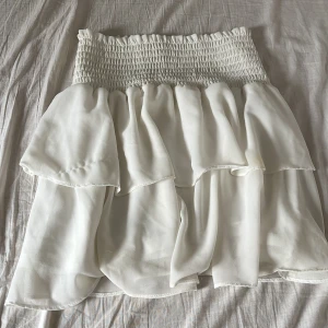 Vit volangkjol från Chelsea - Supersöt vit kortkjol från Chelsea, strechig i midjan och flera lager volanger. Kjolen är luftig och perfekt för varma dagar. Tillverkad i ett lätt, syntetiskt tyg som gör den extra skön. tyvärr har den en fläck men märks inte mycket😔❤️❤️💕 kontakta för fler frågor