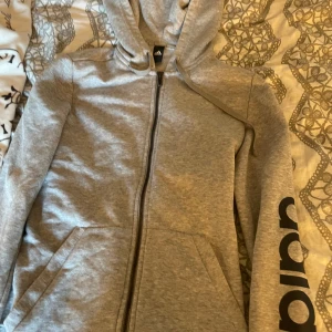 Grå hoodie från Adidas med dragkedja - Snygg grå hoodie från Adidas med hel dragkedja framtill, huva med snörning och fickor på sidorna. Ärmen har svart Adidas-tryck. Perfekt för chill eller träning.