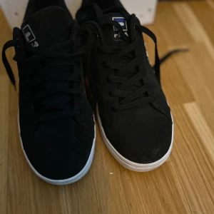 Svarta Fila sneakers med vit sula - Svarta Fila sneakers med klassisk låg profil och vit platt sula. Skorna har snörning och Fila-logga på sidan, plösen och hälen. Ovandelen är i syntetmaterial med en stilren och enkel design som passar till det mesta. Mycket bra skick 