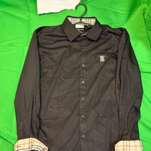 Svart Burberry skjorta med logga - Svart skjorta från Burberry med klassisk krage, knappar framtill och vit broderad TB-logga på bröstet. Insidan av kragen och ärmsluten har det ikoniska rutiga Burberry-mönstret. Skjortan är långärmad och har en stilren, tidlös look.Liknande modell som originalet!