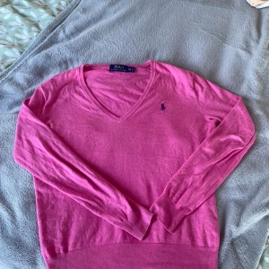 Rosa v-ringad tröja Polo Ralph Lauren - Rosa v-ringad tröja från Polo Ralph Lauren med broderad logga på bröstet. Långärmad och i mjukt material som känns skönt mot huden. Perfekt för dig som gillar klassisk men ändå färgstark stil.