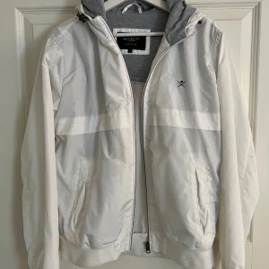 Vit vindjacka från Hackett London S - Snygg vit vindjacka från Hackett London Heritage med grå huva och dragkedja framtill. Jackan har två fickor med lock, ribbade muddar och diskret logga på bröstet. Perfekt för dig som gillar stilren och sportig look.