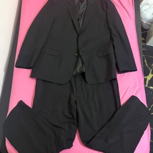 Svart kostym från Savane - Klassisk svart kostym från Savane med enkelknäppt kavaj och matchande byxor. Kavajen har två fickor med lock och en bröstficka, samt knappar vid ärmslut. Kostymen är helsvart och har en stilren, tidlös design som passar perfekt till mer uppklädda tillfällen.