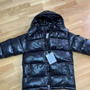 Svart Moncler Maya  - Snygg svart dunjacka från Moncler med glansig finish, huva och dragkedja framtill. Jackan har två fickor med dragkedja och klassisk Moncler-logga på armen. Perfekt för kalla dagar och riktigt stilren look.