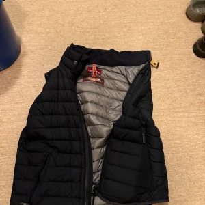 Svart dunväst från Parajumpers - Snygg svart dunväst från Parajumpers med quiltad design och grå insida. Västen har dragkedja framtill, två sidofickor med dragkedja och klassisk Parajumpers-logga i nacken. Perfekt för lager-på-lager och stilren look.