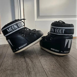 Svarta Moon Boot storlek 36/38 - Moon Boot i svart med vit sula och tydlig logga runt skaftet. Skorna har snörning framtill och är vadderade för extra värme. Materialet är syntet med quiltade detaljer och insidan är mjuk och ljus. Perfekta för kalla vinterdagar och snö.
