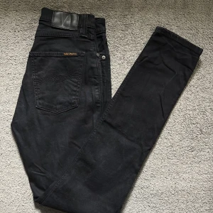 Svarta jeans från Nudie Jeans Co W27 L32 - Svarta jeans från Nudie Jeans Co i modellen W27 L32. Tillverkade av 100% ekologisk bomull med klassisk femficksdesign, svarta sömmar och läderpatch bak. Snygg, rak passform och diskret broderad bakficka. Perfekta för en clean och stilren look.