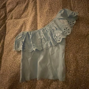 Ljusblå offshoulder topp med volang - Säljer en ljusblå offshoulder topp med bred volang och broderade hålmönster längs kanten. Toppen har en asymmetrisk design med en axel bar och är tillverkad i mjukt bomullstyg. Perfekt för dig som gillar romantiska detaljer och somrig stil.