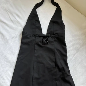 Svart halterneck kortklänning - Svart kortklänning med djup v-ringning och halterneck. Klänningen har en liten rosett och cut-out detalj framtill. Snygg och stilren modell som sitter tight upptill och har en slät finish. Perfekt för dig som gillar minimalistisk och trendig stil. Från SHEIN