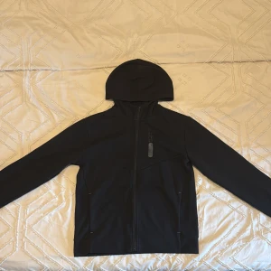 Svart munkjacka - Svart vindjacka med hel dragkedja och huva. Jackan har en stilren design med långa ärmar och två sidofickor. Materialet känns lätt och smidigt, perfekt för blåsiga dagar. Passformen är normal och jackan har en modern look som funkar till streetwear.