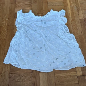 Vit blus med broderade detaljer H&M - Säljer en söt vit blus från H&M med broderade hålmönster och volangkant vid axlarna. Blusen är ärmlös och har en luftig passform, perfekt för varma dagar. Materialet känns lätt och mjukt mot huden. Superfin till jeans eller kjol!