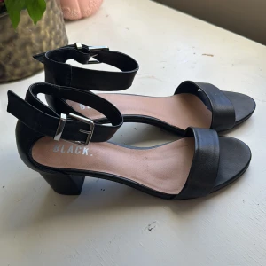 Svarta sandaler med klack och spänne - Stilrena svarta sandaler från BLACK. med bred rem över tårna och justerbart spänne runt ankeln. Skorna har öppen tå, blockklack och är tillverkade i syntetmaterial med en mjuk innersula. Perfekta för dig som gillar enkel och klassisk design. Dessa skor är SÅ bekväma 