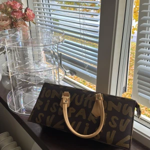  handväska med grafiskt tryck - Cool handväska i brunt med beige handtag och grafiskt mönster i gult. Väskan har två handtag och dragkedja upptill. Perfekt för dig som vill sticka ut med en unik accessoar.