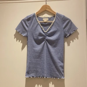 Randig topp från Holly & Whyte - Söt blå- och vitrandig topp från Holly & Whyte med v-ringning och volangkant i spets. Toppen har korta ärmar och en lätt rynkning framtill. Perfekt för dig som gillar en feminin och fräsch stil.