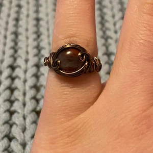 Handgjord ring med röd sten - Unik handgjord ring med en rund, röd sten i mitten och detaljerad trådslingsdesign i bronsfärgad metall. Ringen har en bohemisk vibe och den snirkliga tråden ger ett coolt och personligt uttryck. Perfekt för dig som gillar statement-smycken med vintagekänsla.