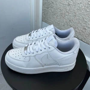 Nike Air Force 1 helvita sneakers - Klassiska Nike Air Force 1 sneakers i helvitt skinn med vit sula och snörning. Ikonisk design med perforerad tå, broderad Nike-logga på hälen och låg profil. Perfekta för dig som gillar en clean och tidlös look.