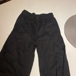 Svarta mjukisbyxor med snörning - Svarta mjukisbyxor med elastisk midja och snörning. Byxorna har en avslappnad passform och är perfekta för chill dagar. Tillverkade i mjukt material som känns skönt mot huden. Enkla att matcha med en hoodie eller t-shirt.