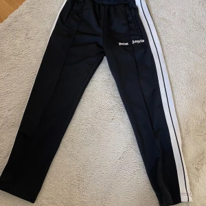 Palm angels mjukis  - Svarta trackpants från Palm Angels med vita sidoränder och logga på vänster ben. Byxorna har elastisk midja och raka ben, tillverkade i polyester för en sportig vibe. Perfekta för en streetwear-look.