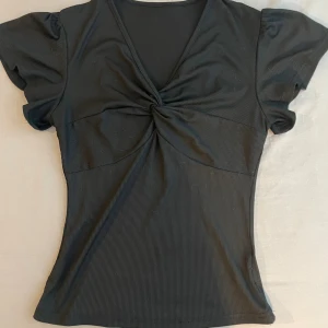 Svart ribbad topp med twistad front - Snygg svart ribbad topp med korta ärmar och twistad detalj framtill. Djup v-ringning ger en trendig look och materialet är stretchigt och följsamt. 