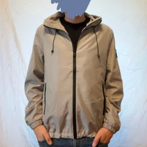 Grå vindjacka med huva och dragkedja - Snygg beige vindjacka med svart dragkedja och huva med snörning. Jackan har elastiska muddar vid ärmslut och nederkant samt två fickor med svarta detaljer. Perfekt för blåsiga dagar och enkel att matcha med olika outfits. Passar M