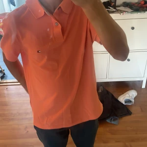 Orange pikétröja Tommy Hilfiger S - Fräsch orange pikétröja från Tommy Hilfiger i storlek S. Klassisk krage, korta ärmar och två knappar framtill. Diskret Hilfiger-logga på bröstet. Materialet är mjuk bomull med regular fit för skön passform. Perfekt för dig som gillar stilrena och färgglada plagg. Köpte för 899 säljer för 599