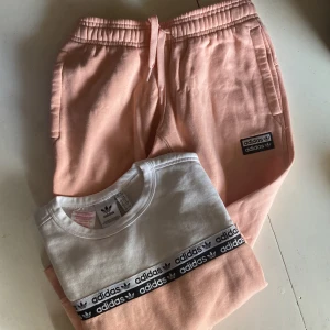 Adidas tracksuit i ljusrosa med vita och svarta detaljer - Säljer en  ljusrosa tracksuit från Adidas med elastisk midja och snörning. Byxorna har muddar med vita ränder vid bensluten och en svartvit Adidas-logga på benet. Swearshirten har loggor ned över armarna och vitt framstycke. Tillverkade i mjuk bomullsmix för maximal komfort.