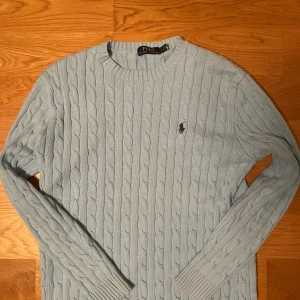 Ljusblå kabelstickad tröja Polo Ralph Lauren - Kabelstickad ljusblå tröja från Polo Ralph Lauren med klassisk rund halsringning och broderad logga på bröstet. Tröjan har långa ärmar och ribbade muddar, perfekt för lager-på-lager under kyliga dagar. Färgen är mer vibrant i verkligheten. Färgen är inte lika klar på bilden pga ljuset.  Inga defekter! Skriv gärna om du har någon fundering. 