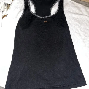 Linne - Svart linnetopp med racerback och diskreta grå detaljer längs kanterna. Toppen har ett litet tryckt logomärke framtill där lite av märket försvunnit därav svårt o tyda vad det är för märke, och ett logomärke på baksidan som har lossnat lite, utöver det inga defekter. Perfekt för träning eller chill dagar.