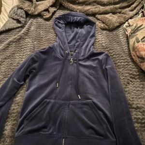 Juicy Couture marinblå velourhoodie XS - Juicy Couture hoodie i mjuk marinblå velour med dragkedja och huva. Klassisk JC-logga broderad på bröstet och snygga metalldetaljer på snörena. Perfekt för en chill och trendig look. Fickor framtill och ikonisk J-dragkedja.