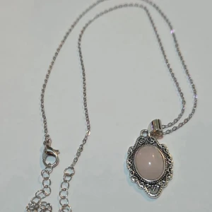Silverhalsband med rosa sten - Ett elegant halsband i silverfärg med tunn kedja och ett detaljerat, vintageinspirerat hänge. Hänget har en oval, ljusrosa sten i mitten omgiven av fina mönster i metall. Perfekt för dig som gillar romantiska och unika smycken.