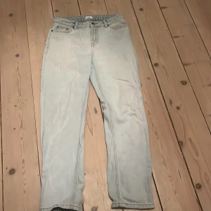 Ljusblå jeans från Grunt - Säljer ett par ljusblå jeans från Grunt med klassisk femficksdesign och raka ben. Jeansen har en snygg tvättad look, silverfärgade knappar och detaljer samt en svart patch bak i midjan. Perfekta för en chill och stilren vibe.