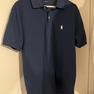 Marinblå pikétröja från Ralph Lauren - Klassisk marinblå pikétröja från Polo Ralph Lauren i custom fit. Tröjan har korta ärmar, krage med två knappar och den ikoniska broderade loggan på bröstet. Säljs pga att den inte passar som önskat. Hör av vid funderingar!