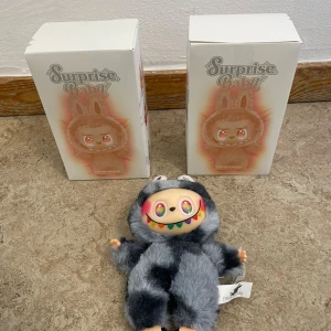 Surprise Baby Plush Doll - Unik Surprise Baby mjukisdocka från The Monsters, levereras med originalförpackning. Dockan har färgglada detaljer och mjuk pälsdräkt. Perfekt skick, inga synliga defekter eller slitage. Samlarobjekt eller present till leksaksentusiasten.