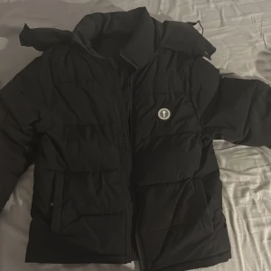 Svart pufferjacka från Trapstar - Trapstar pufferjacka i svart med stor logga broderad på ryggen och rund patch på bröstet. Jackan har huva med tryckknappar, dragkedja framtill och är tillverkad i 100% polyester. Perfekt för kalla dagar och har en riktigt clean streetwear-vibe.