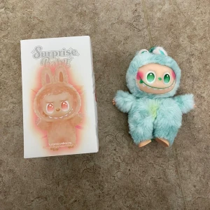 Surprise Baby Plush Pendant Blind Box - Söt Surprise Baby-figur i mjuk plyschdräkt, modell 'Hope' i blått. Kommer med originalförpackning. Perfekt som samlarobjekt eller present. Figuren är i mycket fint skick utan synligt slitage eller defekter.