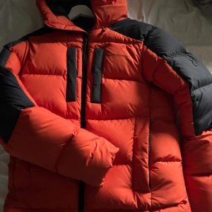 Peak Performance orange pufferjacka - Säljer en fet orange pufferjacka från Peak Performance med svarta detaljer på axlar, ärmar och huva. Jackan har två vertikala bröstfickor med dragkedja, stor huva och markerade sömmar. Perfekt för kalla dagar och riktigt snygg streetstil.