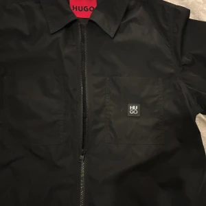 Svart overshirt från Hugo med dragkedja - Stilren svart overshirt från Hugo med klassisk krage, två bröstfickor och diskret logga på vänster sida. Jackan har hel dragkedja framtill och är tillverkad i ett lätt, slätt material som ger en clean look. Perfekt för lager-på-lager och enkel att matcha.